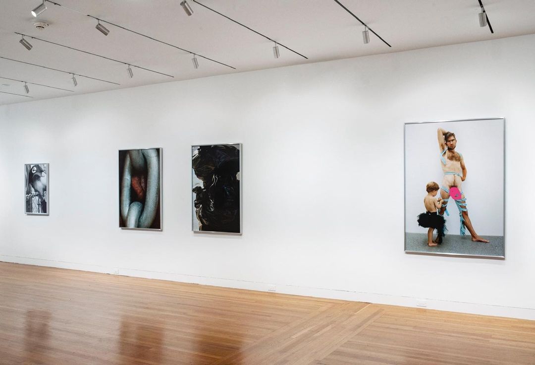 Art gallery featuring framed contemporary photography including a man and child in tutus, displayed on a white wall over a wood floor.