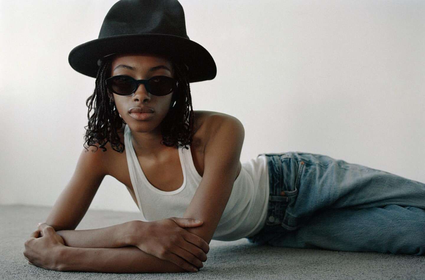 Black woman in a white tank top and black hat lying on a grey carpeted floor wearing sunglasses.