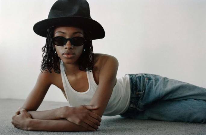 Black woman in a white tank top and black hat lying on a grey carpeted floor wearing sunglasses.