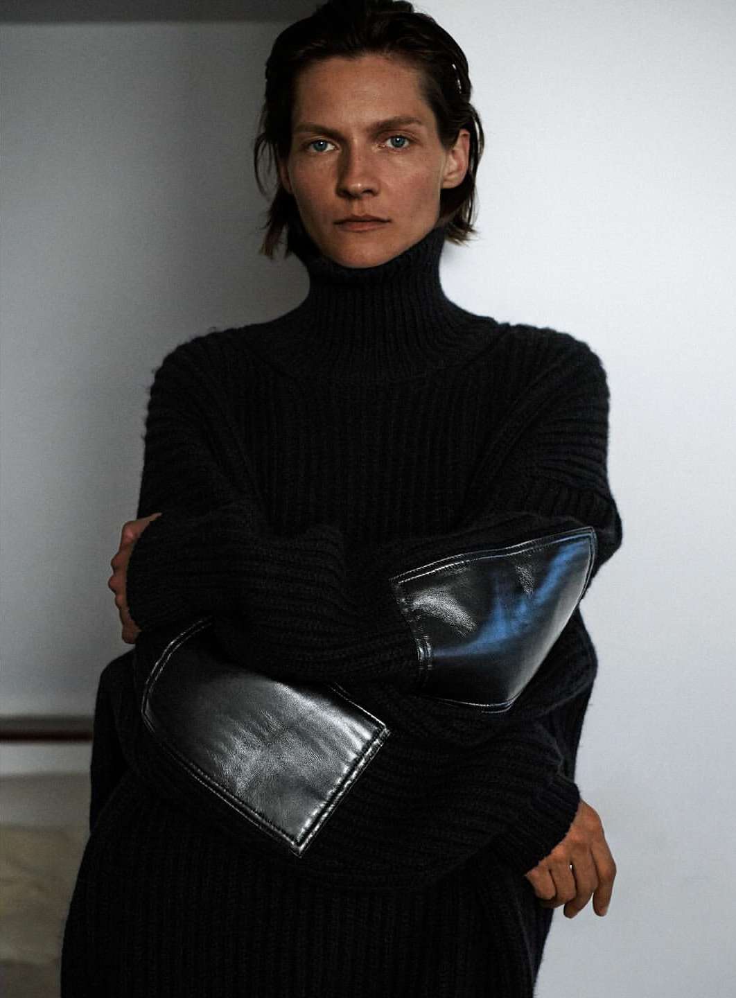 A woman with short hair poses in a black turtleneck sweater with leather elbow patches against a neutral backdrop.