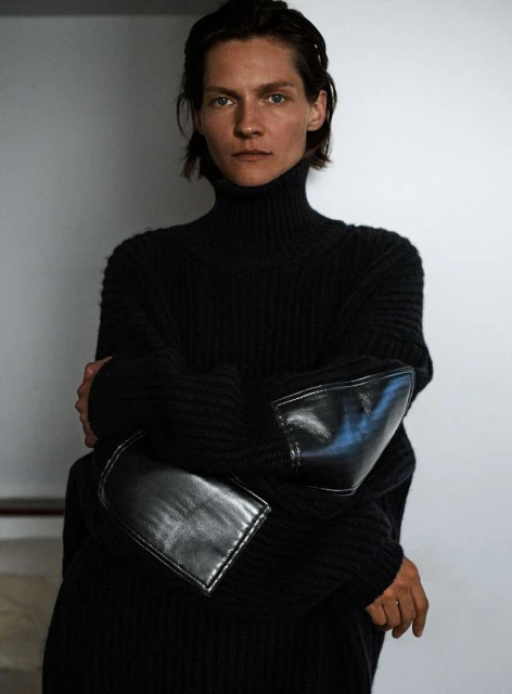 A woman with short hair poses in a black turtleneck sweater with leather elbow patches against a neutral backdrop.