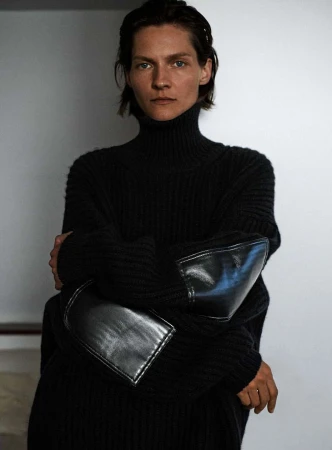 A woman with short hair poses in a black turtleneck sweater with leather elbow patches against a neutral backdrop.