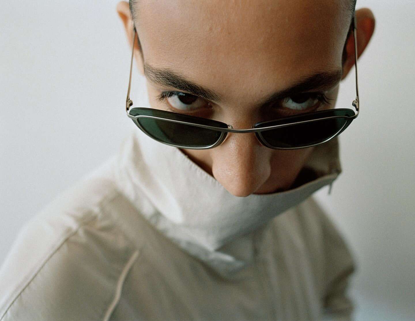 High angle, close-up studio portrait of a young adult wearing sunglasses and a high-collared white shirt, looking intensely at the camera.