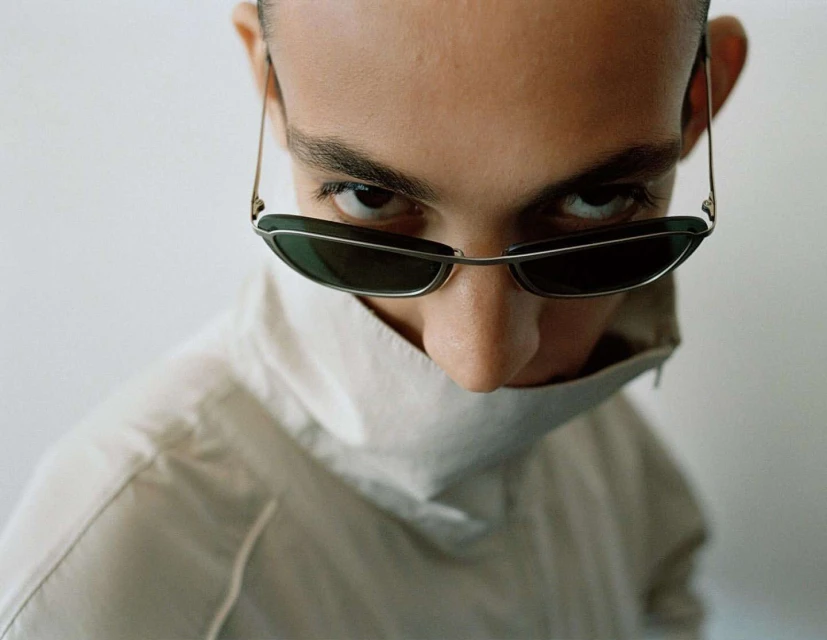 High angle, close-up studio portrait of a young adult wearing sunglasses and a high-collared white shirt, looking intensely at the camera.