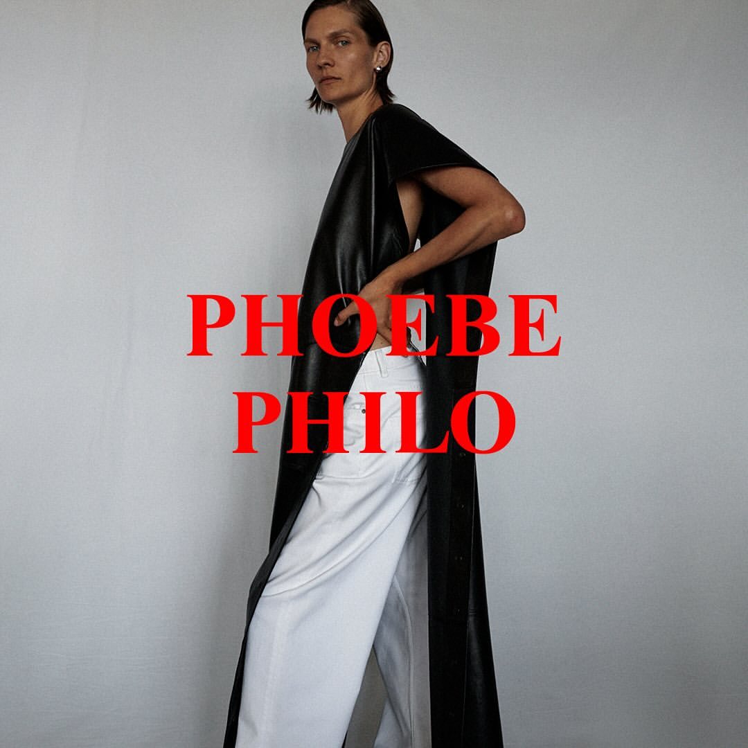 Elegant woman in a draped black tunic and white pants posing against a gray background, fashion-forward and minimalist aesthetic.