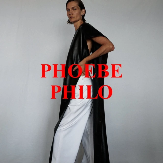 Elegant woman in a draped black tunic and white pants posing against a gray background, fashion-forward and minimalist aesthetic.