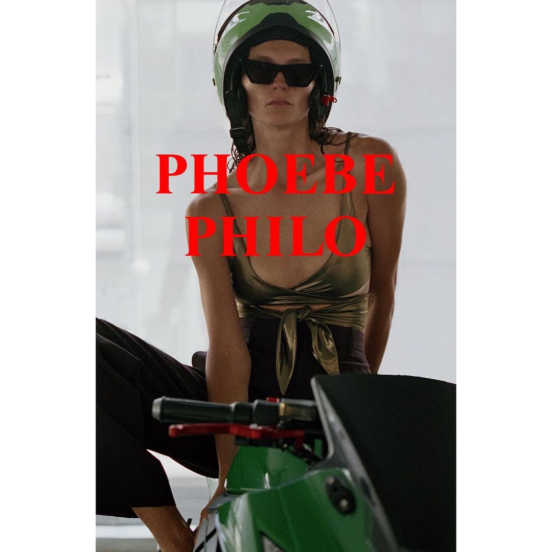 Woman with a helmet sits astride a motorcycle indoors, wearing sunglasses and casual clothes, with red text overlay in a fashion style.