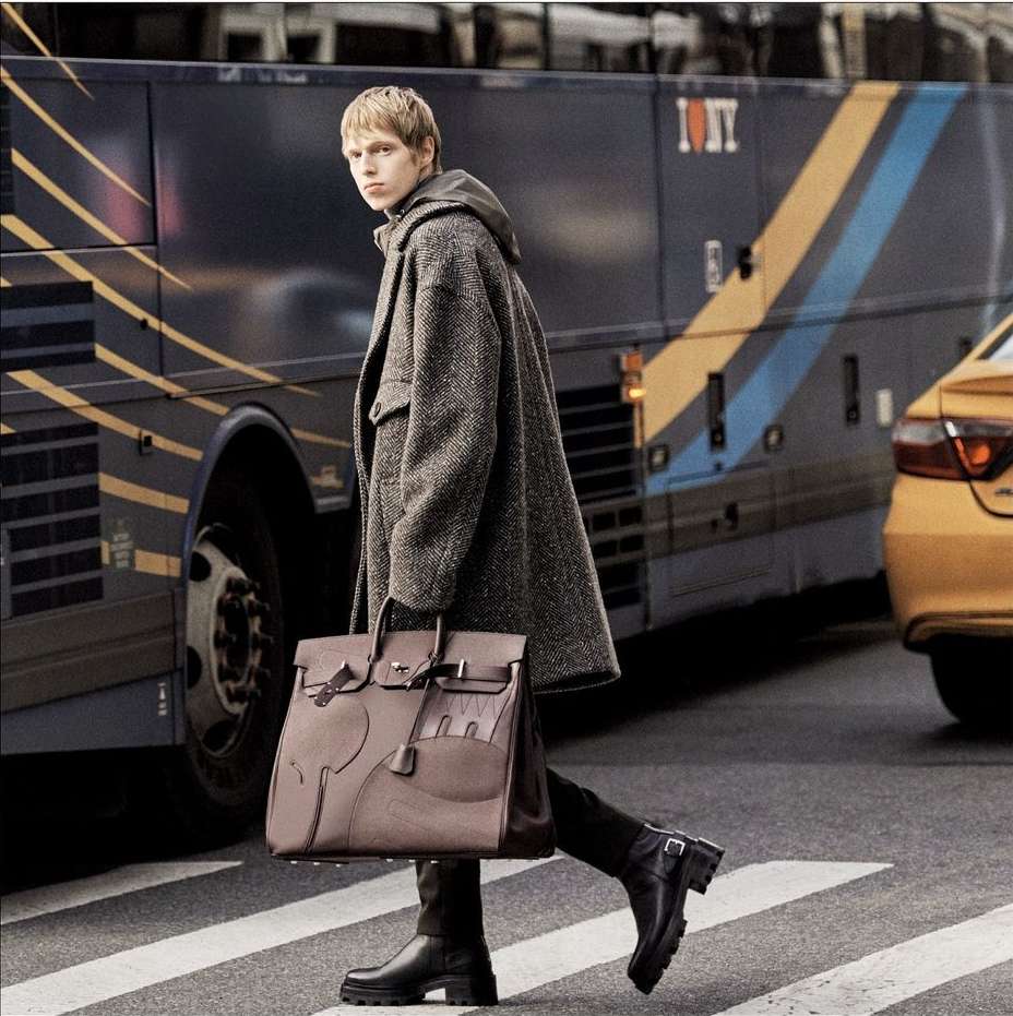 Stylish man walks across a city crosswalk in a herringbone overcoat, holding a structured leather satchel, with a taxi and bus in the background.