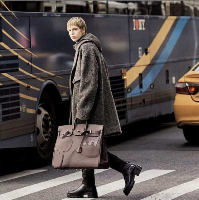 Stylish man walks across a city crosswalk in a herringbone overcoat, holding a structured leather satchel, with a taxi and bus in the background.