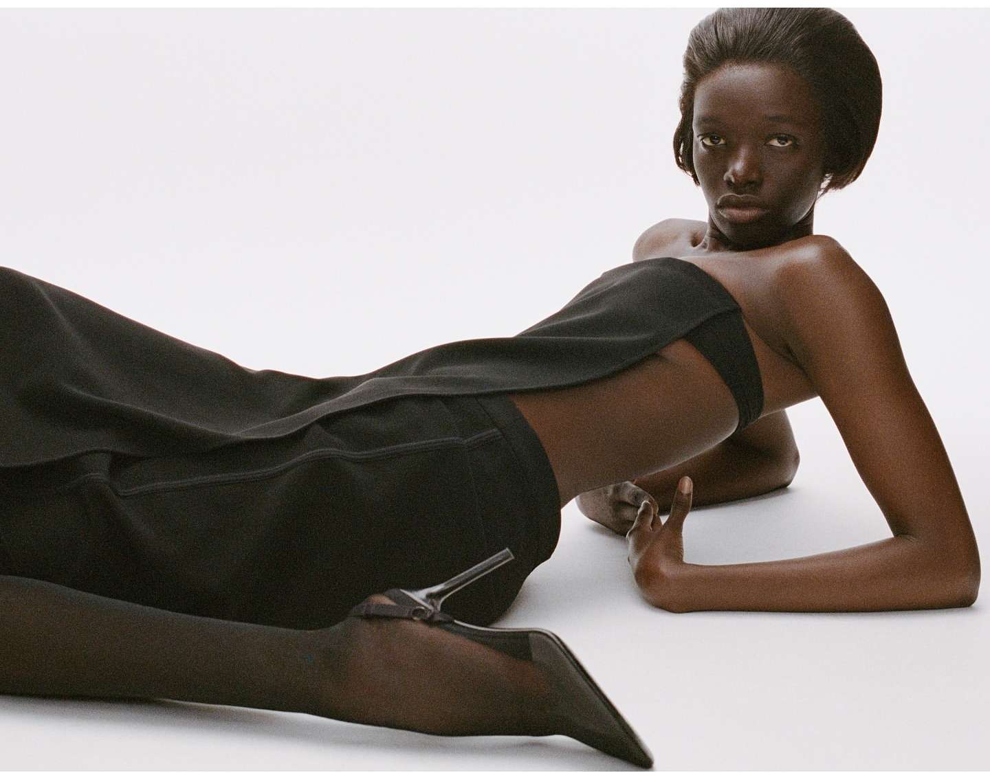 Black woman wearing a strapless black top and matching trousers reclining against a clean white backdrop