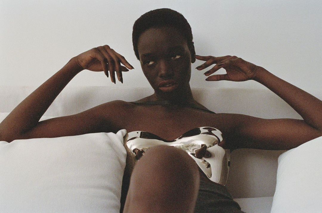 Dark-skinned model poses on a sofa with a reflective gold bustier top against a white backdrop in a fashion or beauty editorial.