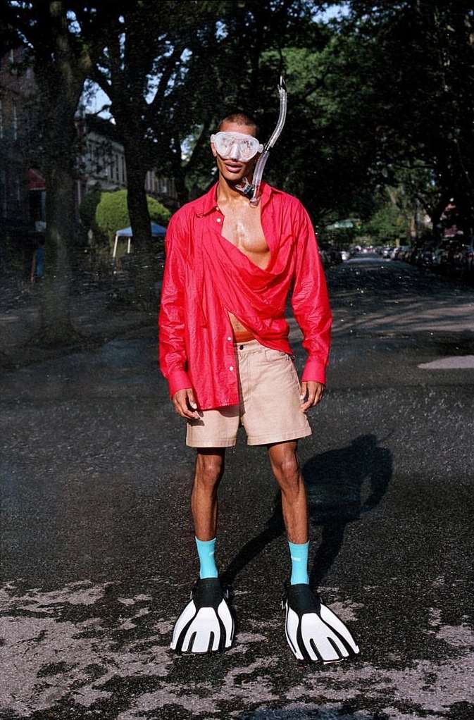 Man in red shirt, shorts, and flippers stands on wet city street with snorkel mask, creating a surreal, playful, and fashion-forward image.