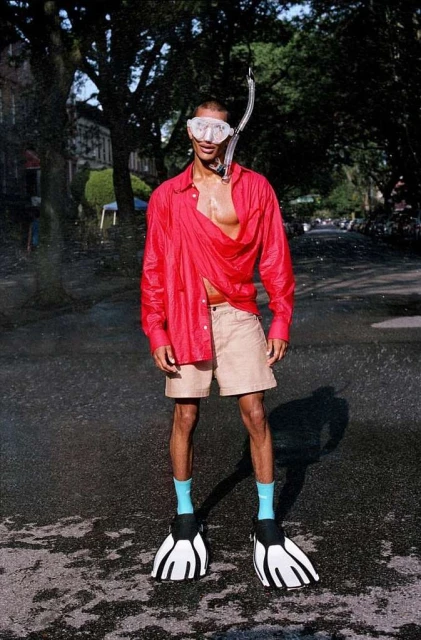 Man in red shirt, shorts, and flippers stands on wet city street with snorkel mask, creating a surreal, playful, and fashion-forward image.