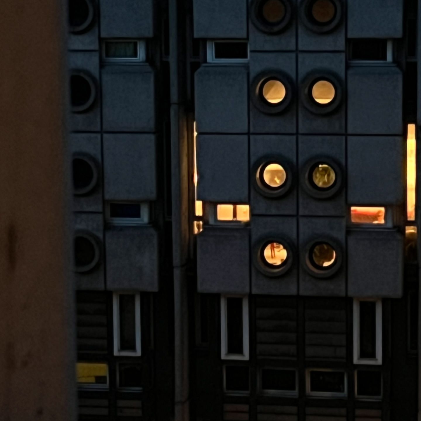 Exterior of a brutalist building at dusk with square windows and illuminated porthole-style window openings, and a grey concrete facade.