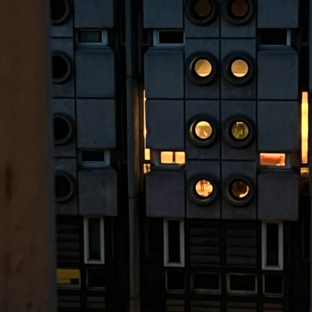 Exterior of a brutalist building at dusk with square windows and illuminated porthole-style window openings, and a grey concrete facade.