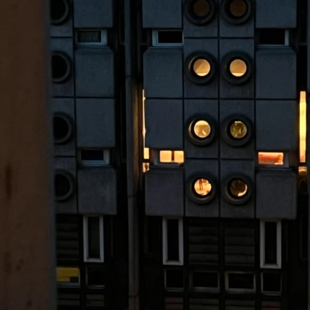 Exterior of a brutalist building at dusk with square windows and illuminated porthole-style window openings, and a grey concrete facade.