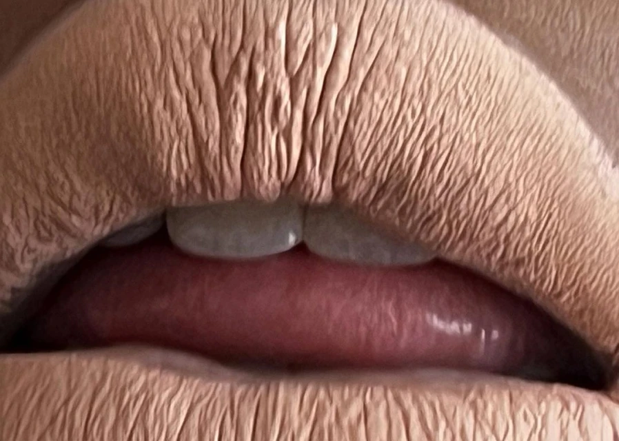 Close-up of a person's mouth featuring beige lipstick on their lips, showing visible teeth and pink tongue.