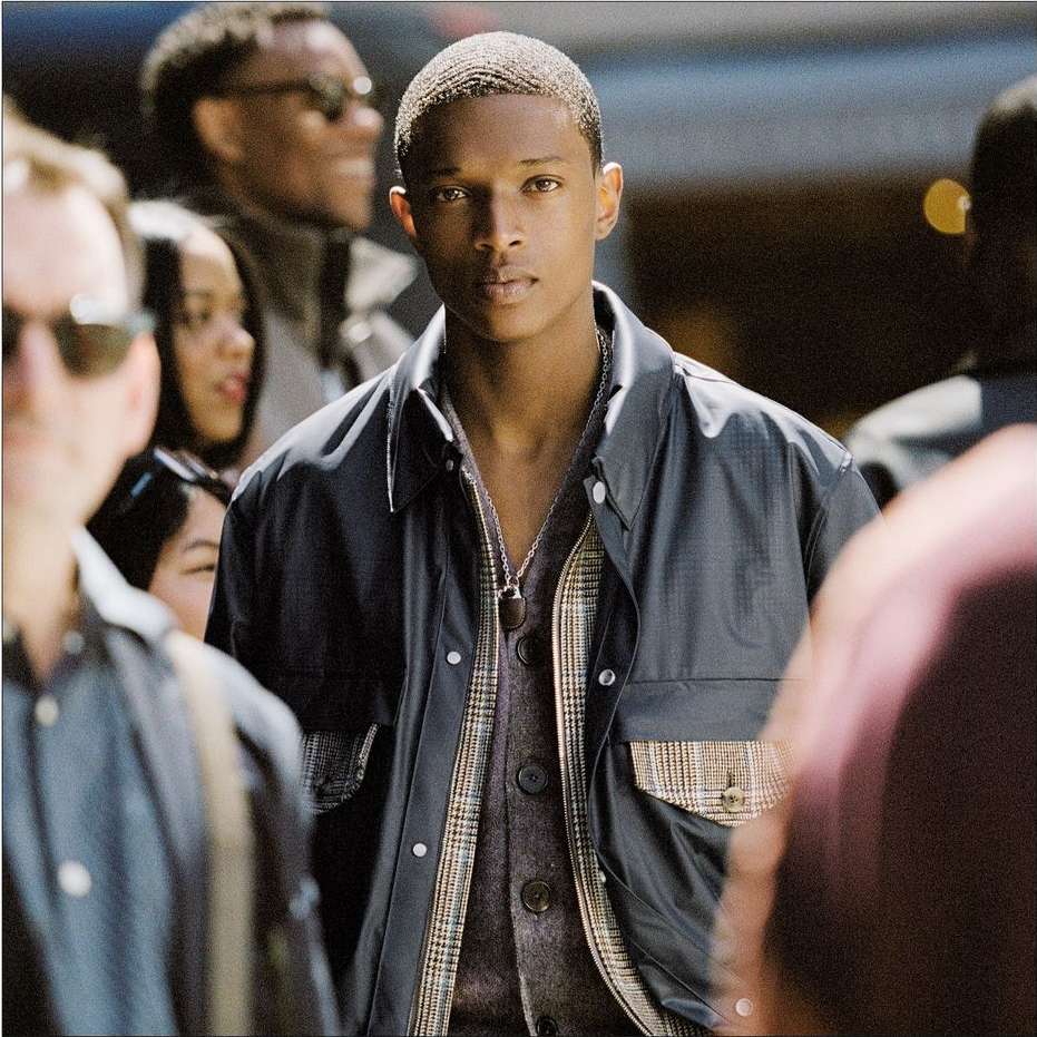 Young Black man with short hair wearing a dark jacket and patterned cardigan amidst a blurred urban crowd