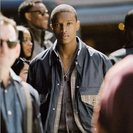 Young Black man with short hair wearing a dark jacket and patterned cardigan amidst a blurred urban crowd