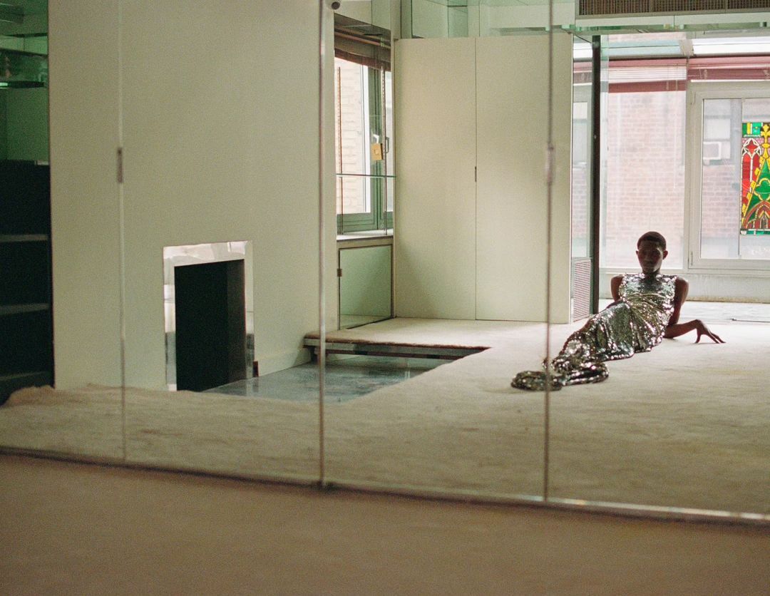 An adult female in a sparkly dress reclines on a light-colored carpet in a mirrored room, creating an illusion of multiple spaces and reflections.