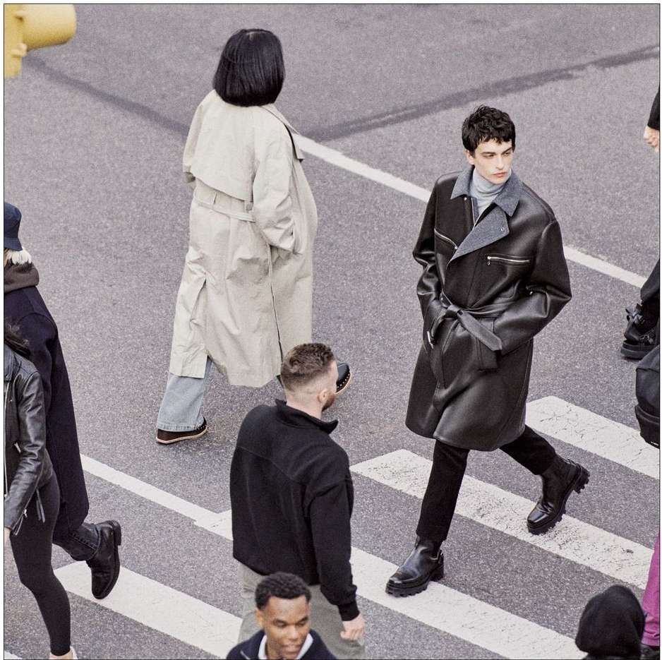 Group of people crossing a crosswalk, one adult male in a dark leather trench coat in the foreground, overcast daylight.