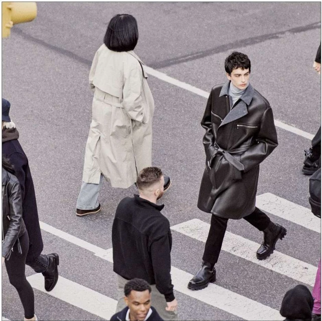 Group of people crossing a crosswalk, one adult male in a dark leather trench coat in the foreground, overcast daylight.