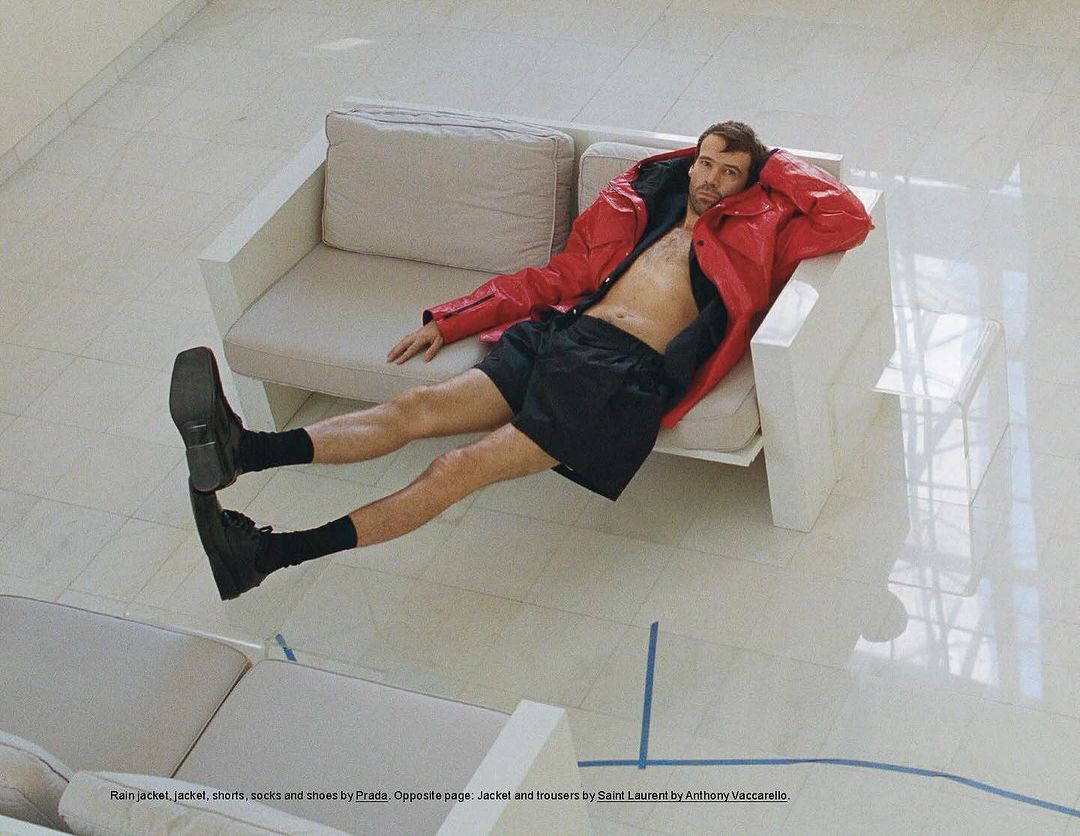 Overhead shot of a man in a red jacket and black shorts reclining on a white couch, with another couch visible below on a tile floor.