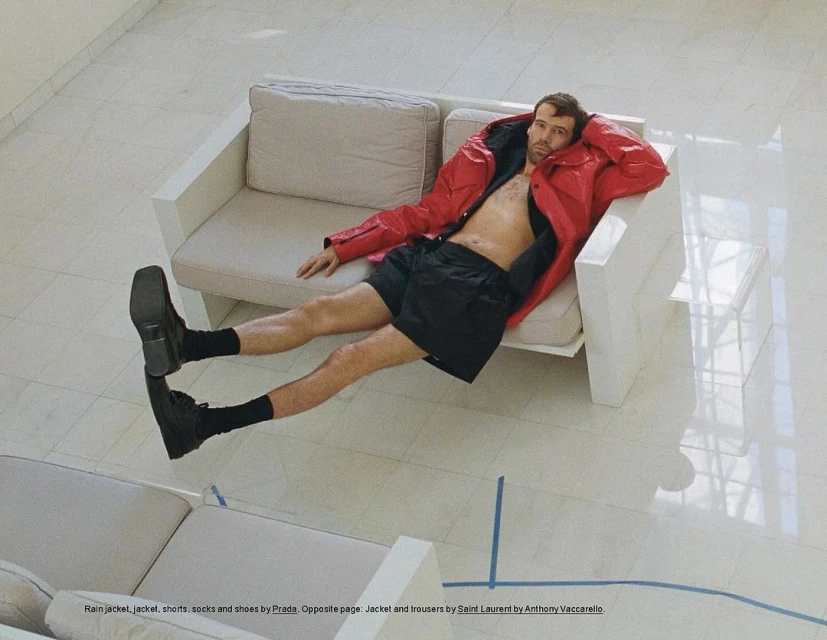 Overhead shot of a man in a red jacket and black shorts reclining on a white couch, with another couch visible below on a tile floor.