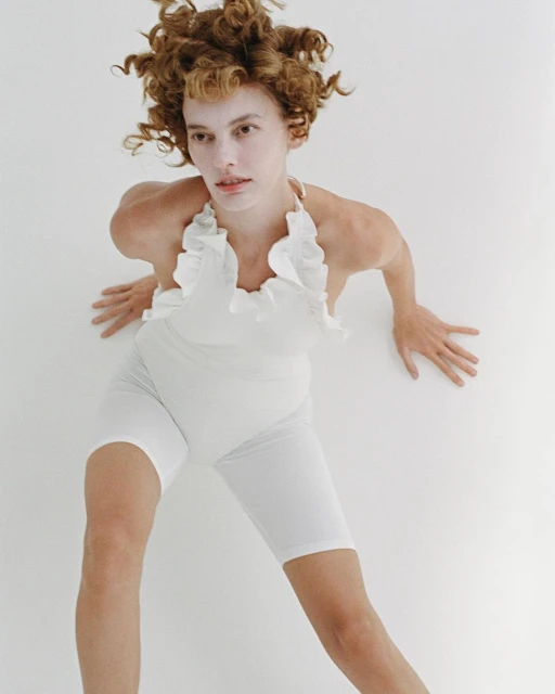 Woman in white ruffled swimsuit and bike shorts posing on white background with curly hair in a high-angle fashion shot.