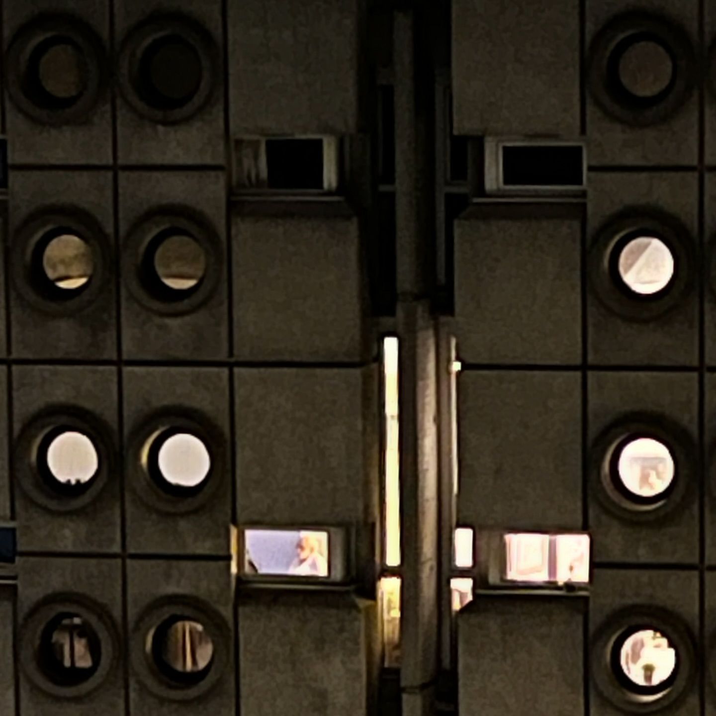 Exterior view of a tall, grey building at night with patterned windows and multiple square window openings with people inside.