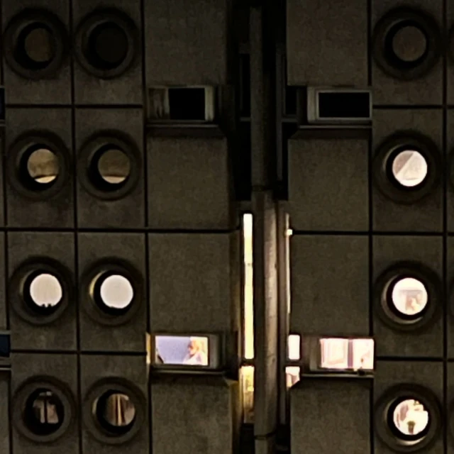 Exterior view of a tall, grey building at night with patterned windows and multiple square window openings with people inside.
