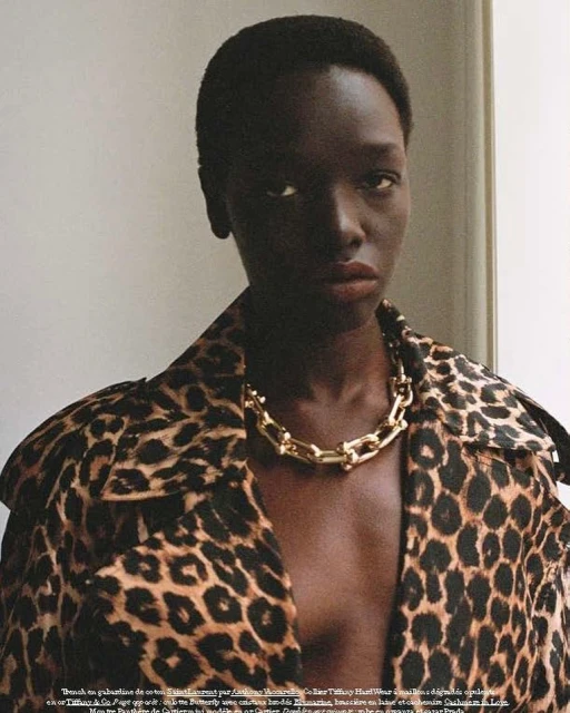 Portrait of a dark-skinned woman wearing an open leopard-print coat and a chunky gold necklace against a neutral background.