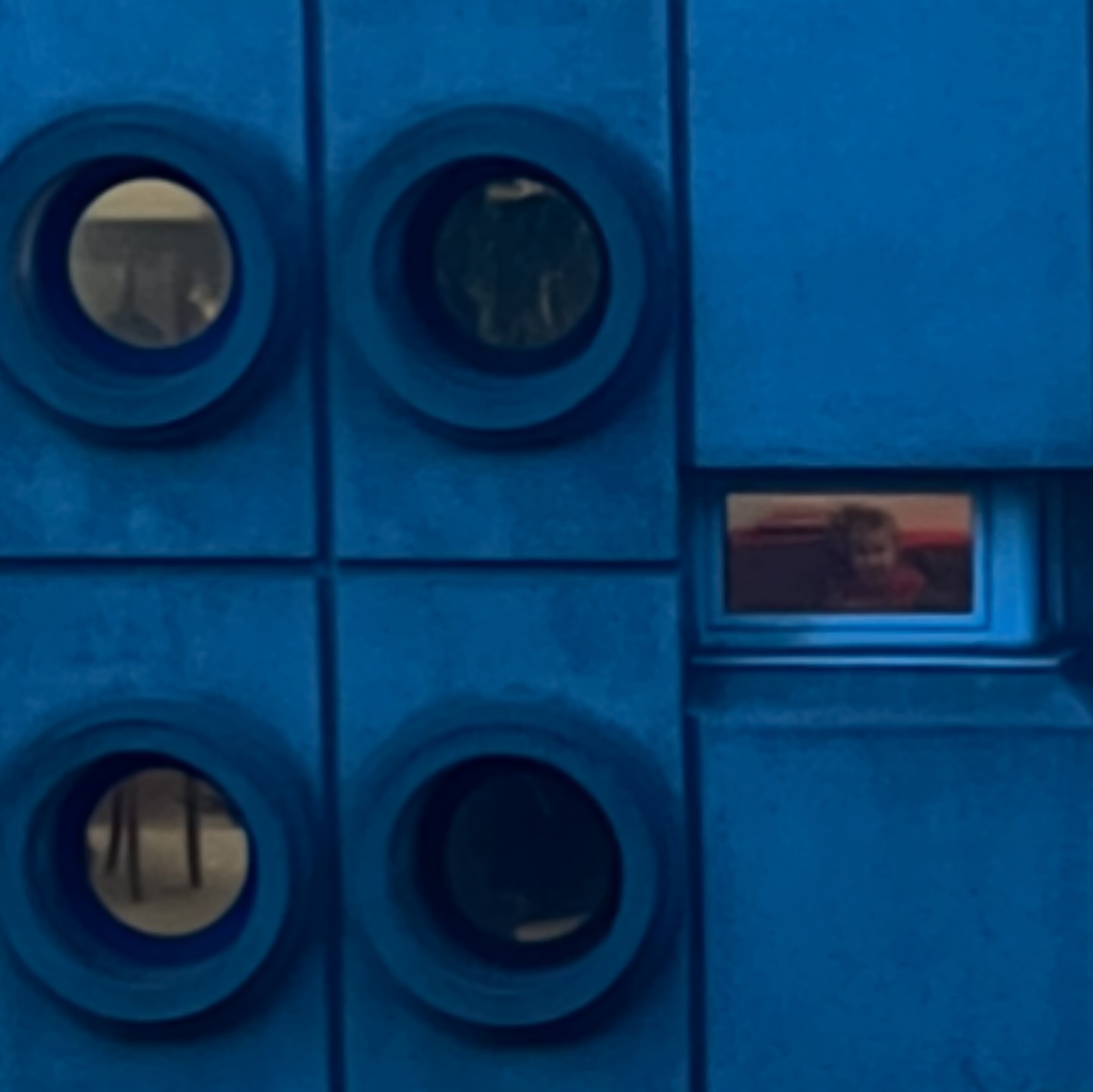 Close-up of a blue Lego brick wall with circular openings and a rectangular opening showing a child looking out from the structure.