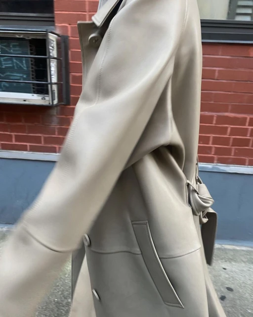 Fashionable person wears a tan trench coat on an urban street with brick building, showcasing style and classic outerwear.