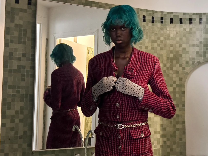 Young Black woman in a pink suit and turquoise wig stands in a tiled bathroom next to a mirror reflecting her image.