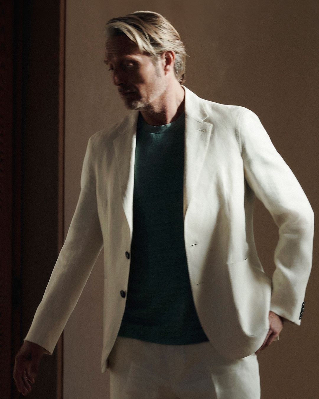 Man with blond hair in a light-colored suit poses indoors in soft light. He is wearing a teal t-shirt and looking off to the side.