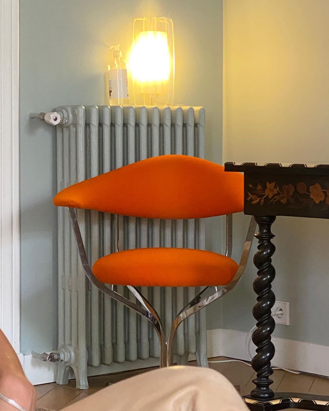 Interior view of an orange mid-century modern chair next to a black table, a vintage radiator and a glowing table lamp.