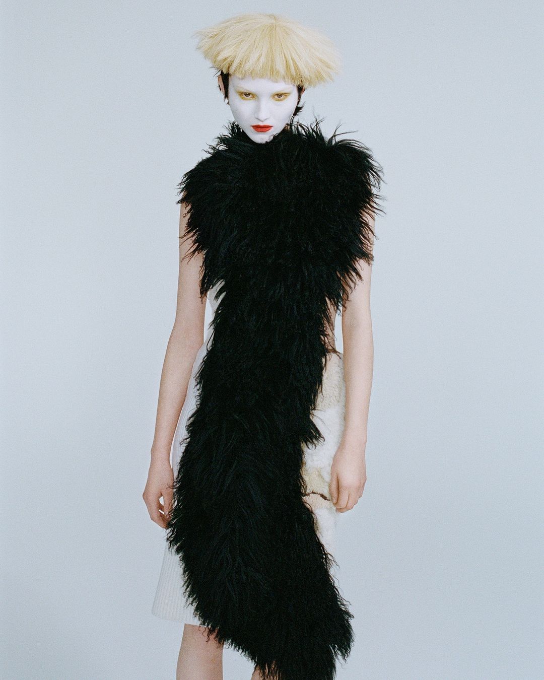 Fashion model with white face paint and unusual blonde wig wears a black feathered shawl, striking a pose against a neutral background.