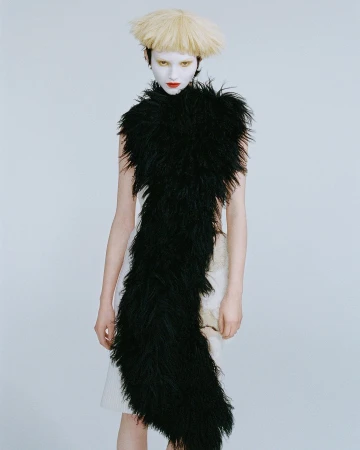 Fashion model with white face paint and unusual blonde wig wears a black feathered shawl, striking a pose against a neutral background.