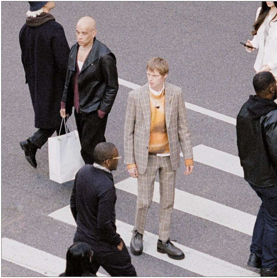 Male fashion model wears plaid suit in a street fashion editorial with people crossing the road on a crosswalk in daylight.