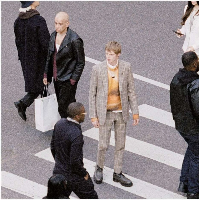 Male fashion model wears plaid suit in a street fashion editorial with people crossing the road on a crosswalk in daylight.