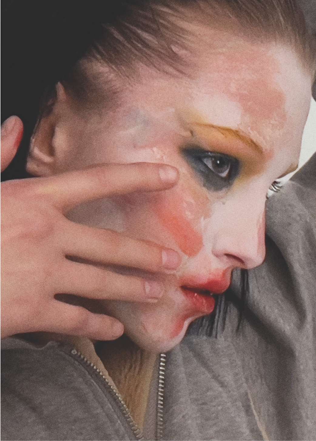 Adult with heavy makeup applies makeup to face; red lips, black eyeshadow, yellow eyebrows, white face makeup and neutral background.