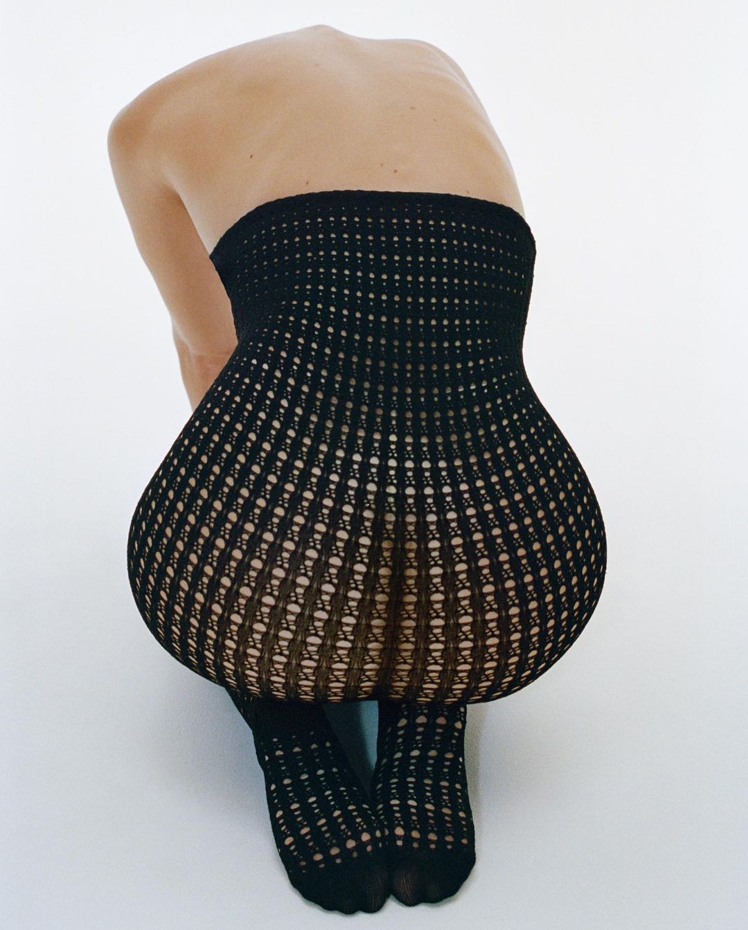 Back view of a person crouching with open-weave black tights, in a white photo studio, showcasing fashion and style.