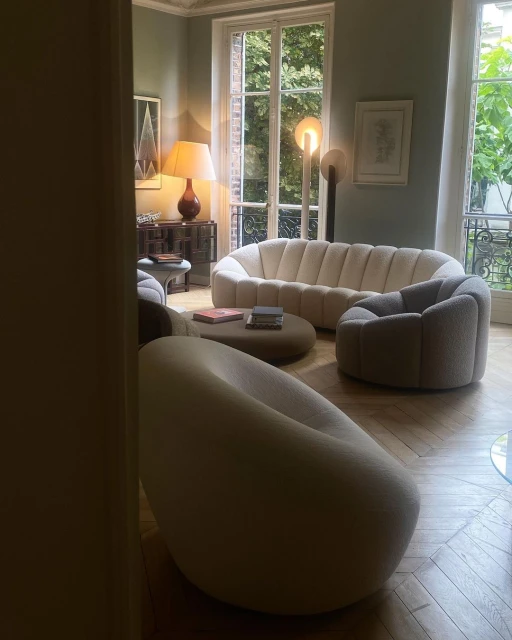 Living room interior with a cream-colored sofa, a round coffee table, and a matching armchair, illuminated by natural light and a table lamp.