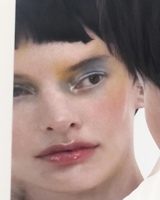 Close-up studio shot of a woman with black hair, blue eyeshadow, and glossy lips, reflected in a mirror.