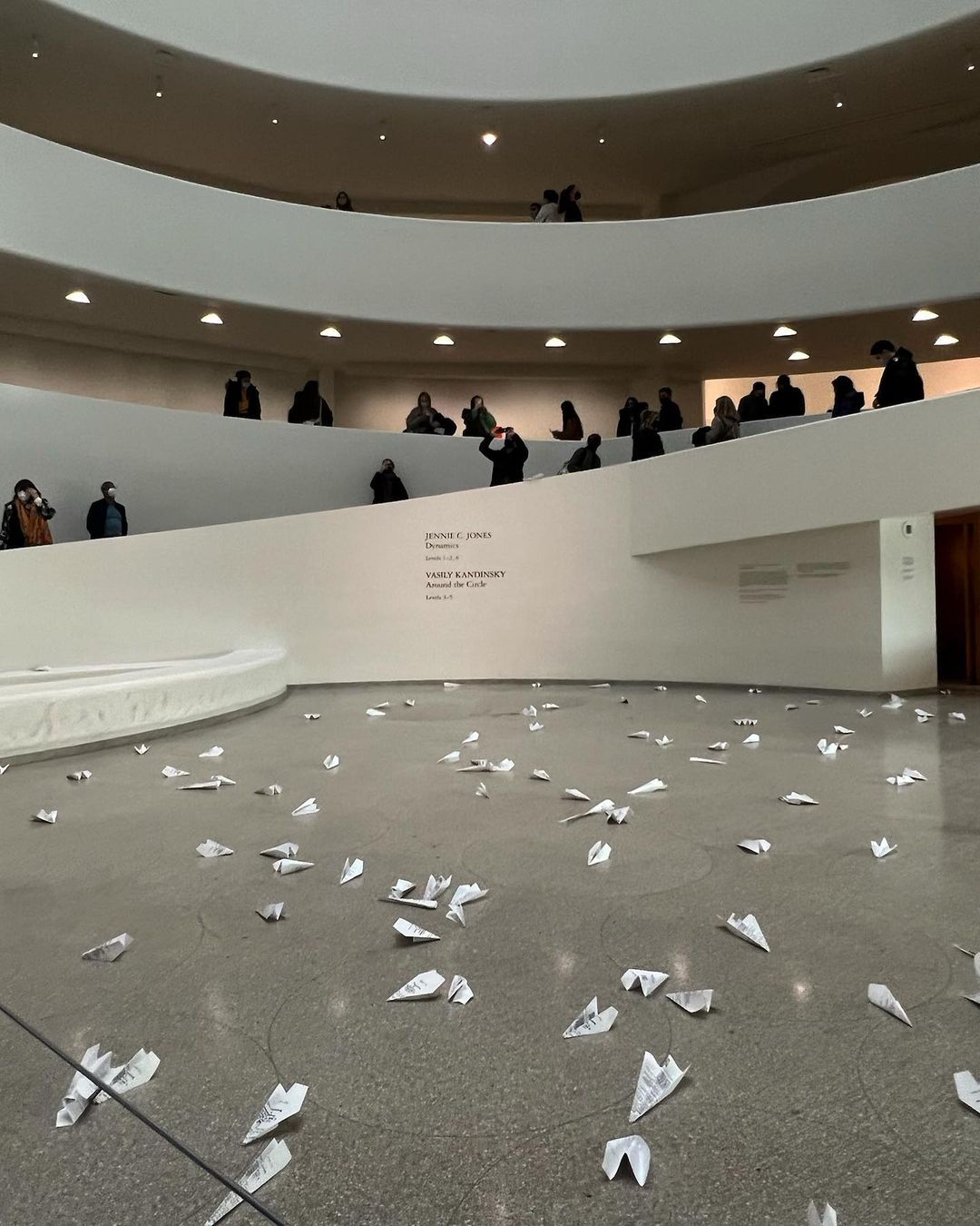 Interior view of the Guggenheim Museum featuring many paper airplanes scattered on the floor and visitors on the spiral ramps above.