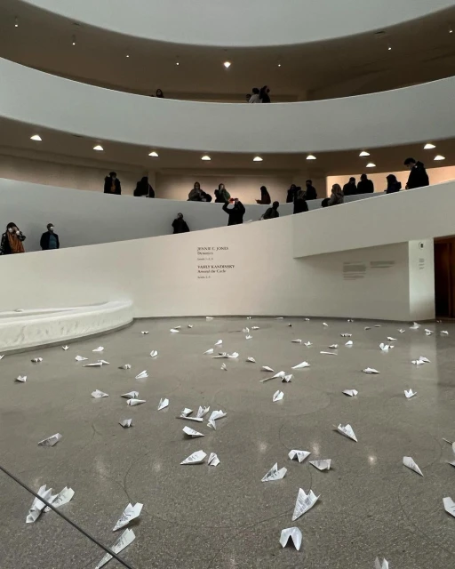Interior view of the Guggenheim Museum featuring many paper airplanes scattered on the floor and visitors on the spiral ramps above.