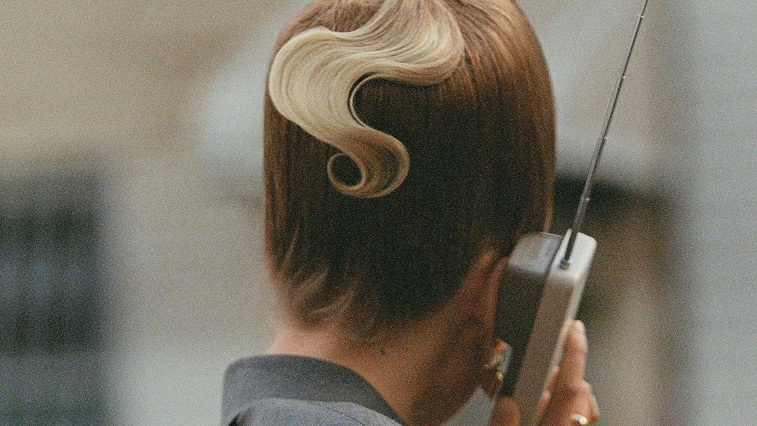Woman with a retro hairstyle talking on a vintage cell phone, viewed from behind with a neutral gray background, close up shot.