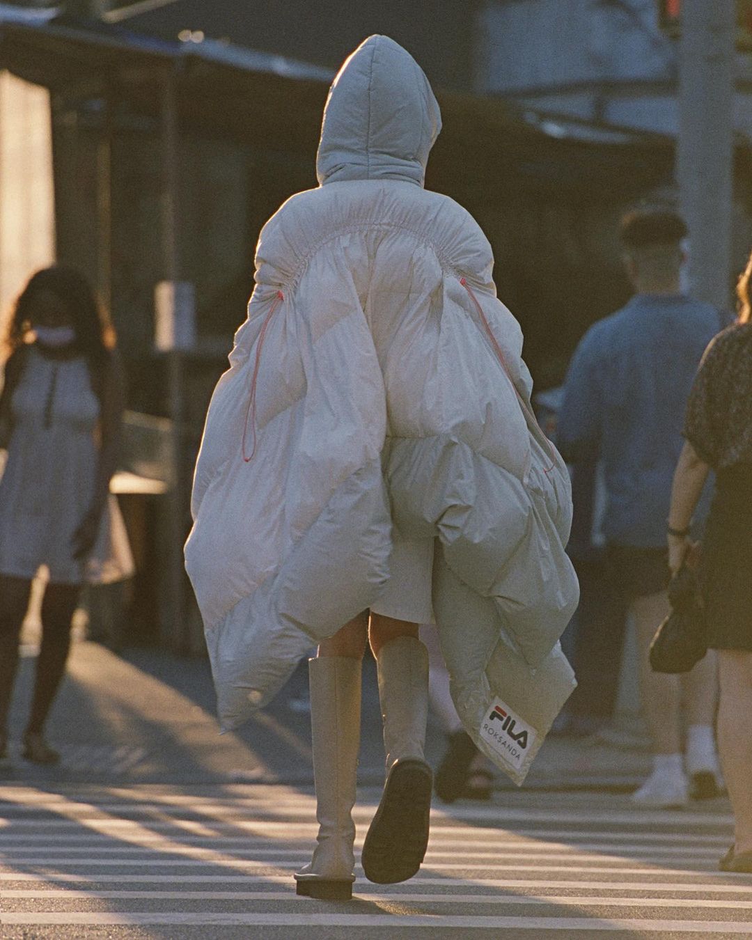 Person wearing oversized white puffer coat crossing a street in an urban setting with other people visible in the background.