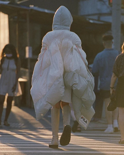 Person wearing oversized white puffer coat crossing a street in an urban setting with other people visible in the background.